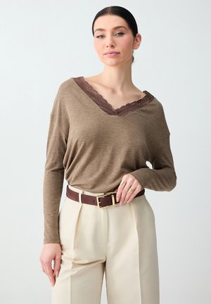 Femme portant un haut marron à manches longues avec col en V et bordure en dentelle, un pantalon crème taille haute et une ceinture marron, posant devant un fond uni.