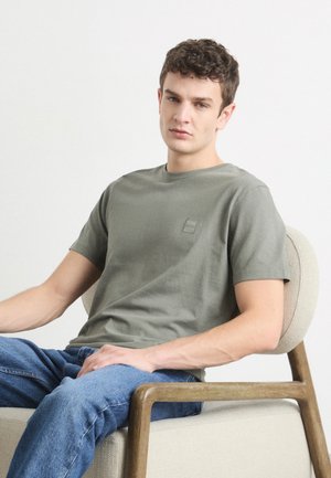 Olivovo zelené tričko s okrúhlym výstrihom a krátkymi rukávmi, so malým štvorcovým logom na hrudi. Nosené s modrými džínsami.