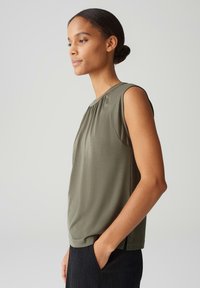 Olive green sleeveless blouse, soft fabric, round neckline with pleats, relaxed fit, side slits, worn with a textured black skirt.