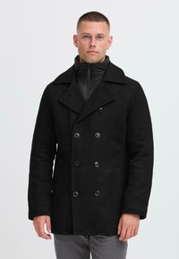 Schwarzer doppelt gefütterter Wollmantel mit großen Knöpfen, breiten Revers und einer abnehmbaren schwarzen Steppjacke im Inneren. Glatte Textur, taillierte Passform.
