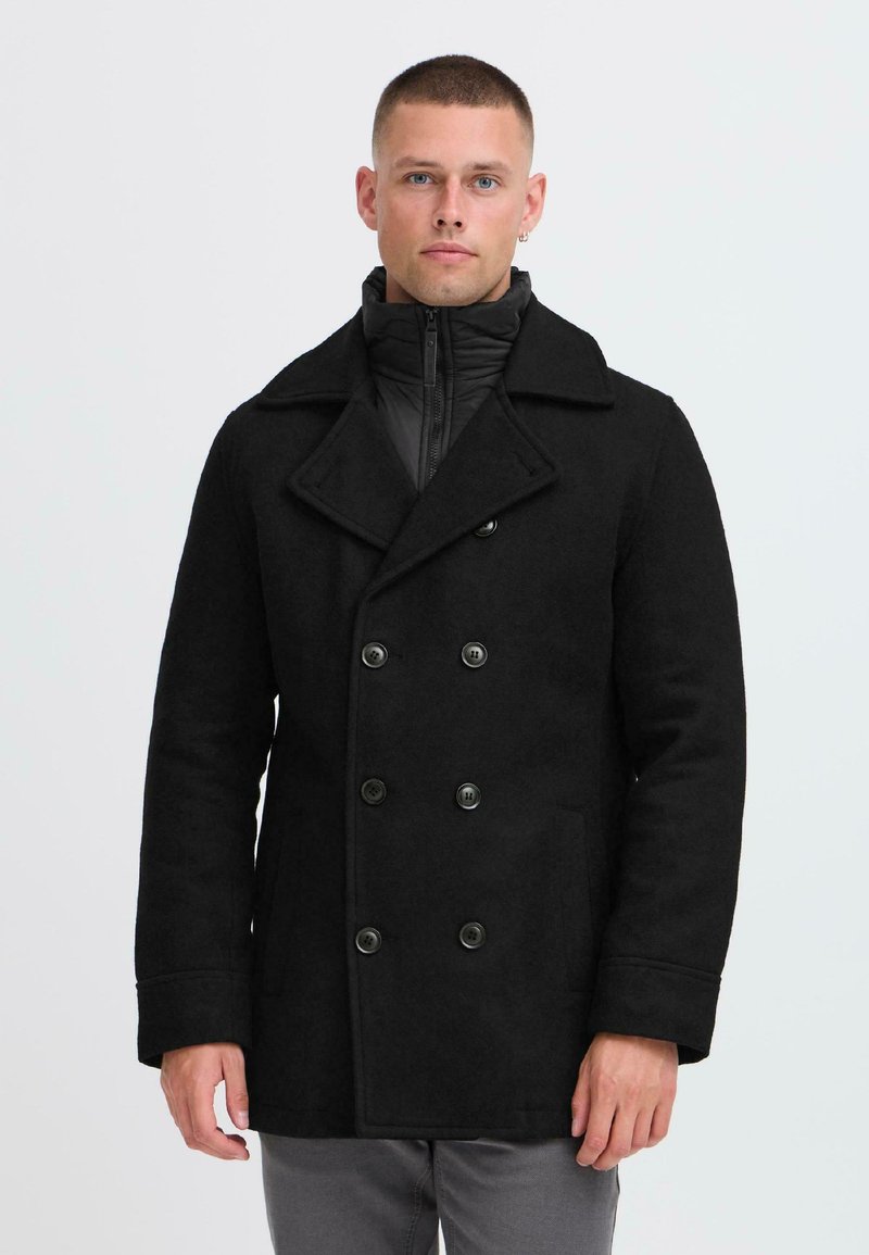 Schwarzer doppelt gefütterter Wollmantel mit großen Knöpfen, breiten Revers und einer abnehmbaren schwarzen Steppjacke im Inneren. Glatte Textur, taillierte Passform.