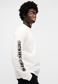 Λευκό μακρυμάνικο πουκάμισο με μαύρο γραφικό «DICKIES CREW» τυπωμένο κάθετα στο αριστερό μανίκι. Βαμβακερό υλικό, άνετη εφαρμογή, μινιμαλιστικός σχεδιασμός.