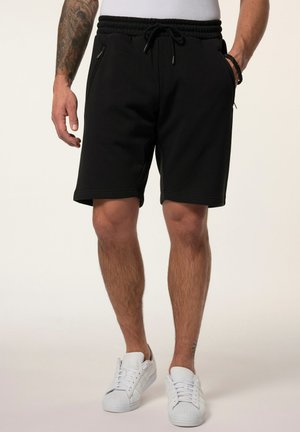 Homme portant un short noir à cordon avec des poches zippées et des baskets blanches, debout devant un fond uni.