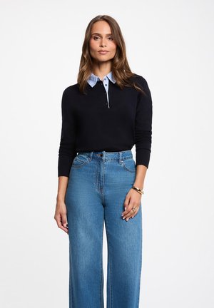 Donna con lunghi capelli castani che indossa una maglia nera a maniche lunghe con colletto azzurro chiaro, jeans blu vita alta, e braccialetto e anelli d'oro.