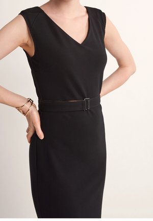 Vrouw die een zwarte mouwloze jurk met V-hals en bijpassende riem draagt, handen op de heupen, gouden geometrische armband aan haar linkerpols.