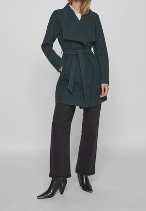 Femme portant un manteau ceinturé vert foncé sur une chemise blanche, associé à un pantalon large noir et des bottines à talons noires.