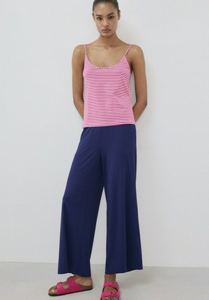 Femme debout, les mains dans le dos, portant un débardeur rayé rose et blanc, un pantalon large bleu marine et des sandales roses avec une boucle devant un mur blanc.