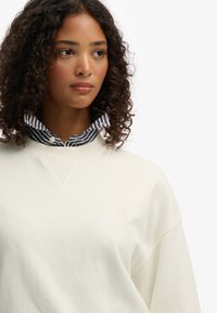 Sudadera de tela suave en color crema con cuello en V. Llevada sobre una camisa de cuello a rayas azul y blanca, mostrando un look casual en capas.