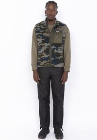 Kamizelka z polaru w kamuflażu w odcieniach zieleni, niebieskiego i szarości, nałożona na oliwkową koszulę z długim rękawem, w zestawie z czarnymi spodniami cargo i ciemnym obuwiem.