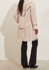 Knit cardigan in pale pink with floral embroidery, fringed hem, and waist tie. Paired with black flared pants and heeled boots.