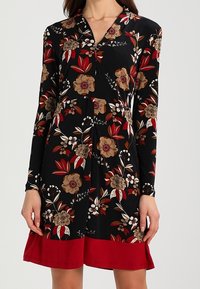 Černé dlouhé šaty s rukávy a hnědým a červeným květinovým potiskem, výstřih do V a jednobarevným červeným lemem, které má na sobě stojící osoba.