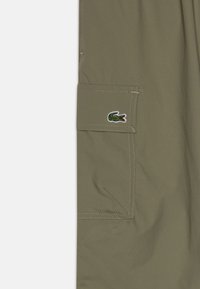 Olive zelene kratke hlače s cargo žepom in kontrastnim šivom. Imajo izvezen logo Lacoste v zeleni in rdeči barvi.