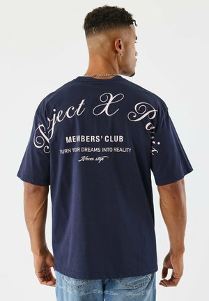 Hombre con cabello rizado corto y arete, vistiendo una camiseta azul marino con "Project X Paris" y texto motivacional en la parte de atrás, combinada con jeans azules.