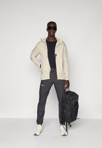 Beige lightweight jacket with hood, black t-shirt, dark gray pants, white sneakers, black backpack, standing pose, sunglasses, minimal background.