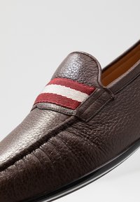 Mocassin en cuir marron avec une surface texturée et un détail cousu, agrémenté d'une bande en tissu rayée rouge et blanche sur le dessus.