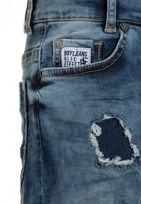 Jeans in denim blu per ragazzi, con una toppa strappata sulla coscia, cuciture a contrasto e un'etichetta "Blue Effect" sulla cintura.