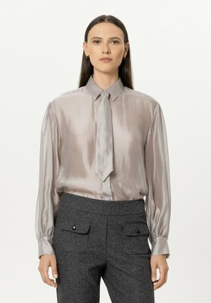Woman with straight dark hair wears shiny silver blouse with matching tie and high-waist dark gray pants with front flap pockets.