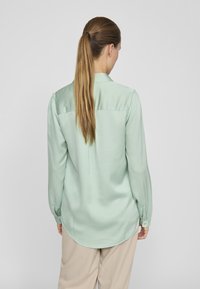 Mint green silk blouse with long sleeves, a collared neck, and buttoned cuffs, featuring a slightly curved hem and smooth texture.