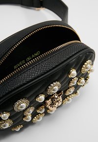 Borsa a vita nera trapuntata con zip dorata, decorazioni in perle e borchie dorate, e un dettaglio centrale di testa di leopardo dorata.