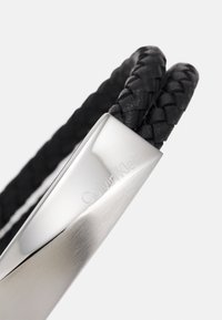 Pulsera de cuero trenzado negro con un cierre de metal plateado pulido grabado con "Calvin Klein". Textura suave y diseño elegante.
