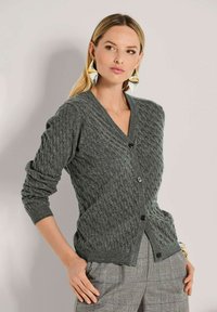 Frau in einem grauen Kabelstrick-Cardigan mit schwarzen Knöpfen, karierten Hosen, großen metallischen geometrischen Ohrringen und einem goldenen Armband, die seitlich posiert.