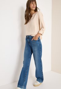 Beige bluse med V-hals og op-rullede ærmer, parret med brede blå jeans. Modellen har lyse sneakers på, hvilket giver et afslappet look.
