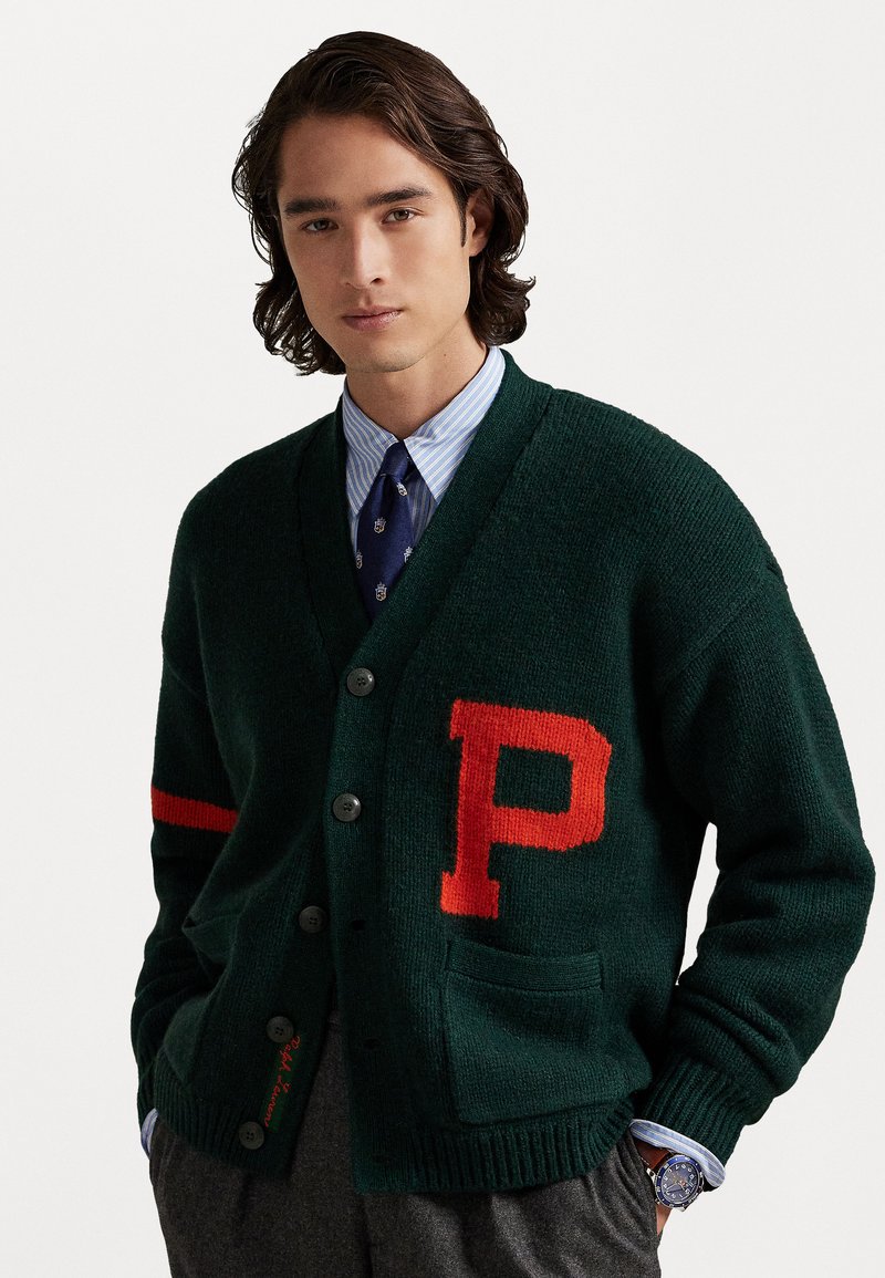 Roheline kudum cardigan punase "P" logoga ja triibulise aktsendiga. Omab kolme nuppu, taskut ja soonikuga varrukaid. Kantud sinise triibulise särgi peal.