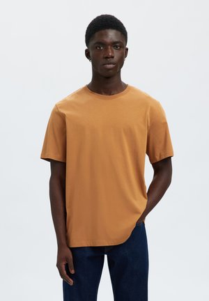 Jeune homme aux cheveux courts portant un t-shirt uni à manches courtes couleur brun clair et un jean bleu foncé, debout avec une main dans la poche.