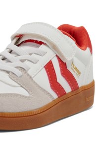 Weißer Sneaker mit roten Akzenten, Materialien aus Wildleder und Leder, Klettverschluss, strukturiertes Gummisohle und "hummel" Branding an der Seite.