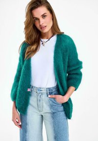 Žena v tyrkysovom mäkkom kardigáne cez biele tričko a džíny s vysokým pásom v dvoch odtieňoch, pózuje s rukou v vrecku na bielom pozadí.