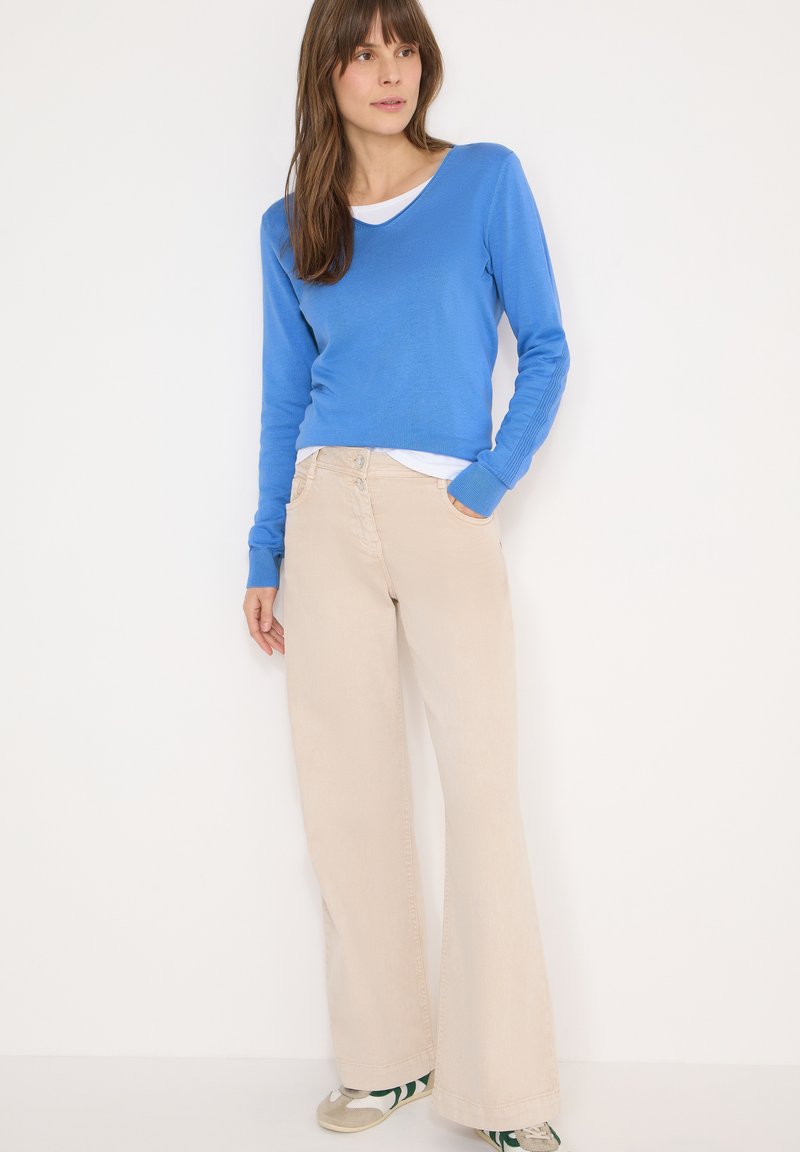 Pull bleu à manches longues avec col en V porté sur une chemise blanche, associé à un pantalon beige à jambes larges et des baskets avec des accents verts.