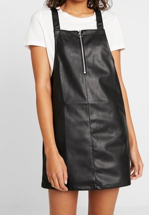 Donna che indossa un vestito salopette nero in ecopelle con cerniera frontale sopra una t-shirt bianca a maniche corte, in piedi davanti a uno sfondo semplice.