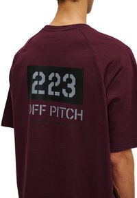 Bordeauxfärgad bomullströja med en svart loppa med vita siffrorna "223" och orden "OFF PITCH" i grått, med koordinater tryckta nedanför.