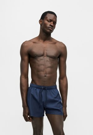 Štíhly svalnatý muž v námornícky modrých plavkách stojaci na jednofarebnom bielom pozadí.