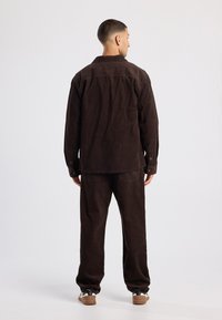 Brown corduroy shirt jacket with buttoned cuffs, paired with matching corduroy pants. Textured fabric and relaxed fit, shown from behind.