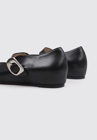 Zapatos de cuero negros con punta redondeada, un detalle de hebilla en el costado y un tacón bajo. El nombre de la marca está sutilmente grabado en el tacón.