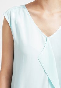 Blusa senza maniche di colore azzurro chiaro con scollatura a V. Presenta un tessuto morbido e un accento drappeggiato nella parte anteriore, che conferisce una silhouette fluida.