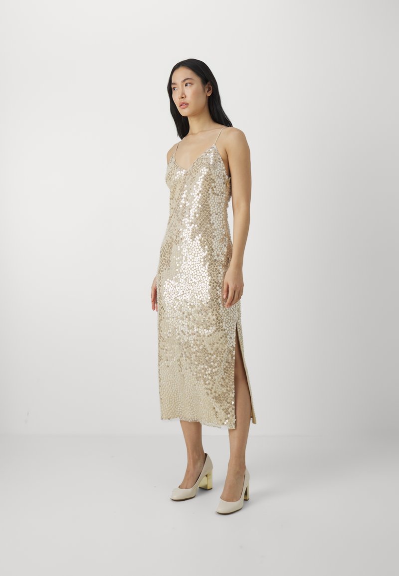 Goldfarbene Pailletten-Midi-Kleid mit einer taillierten Silhouette, Spaghettiträgern und einem seitlichen Schlitz, kombiniert mit cremafarbenen Blockabsatzpumps.