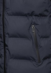 Veste matelassée bleu marine avec une finition mate, dotée d'une poche zippée à gauche avec une fermeture éclair noire élégante et des sections matelassées.