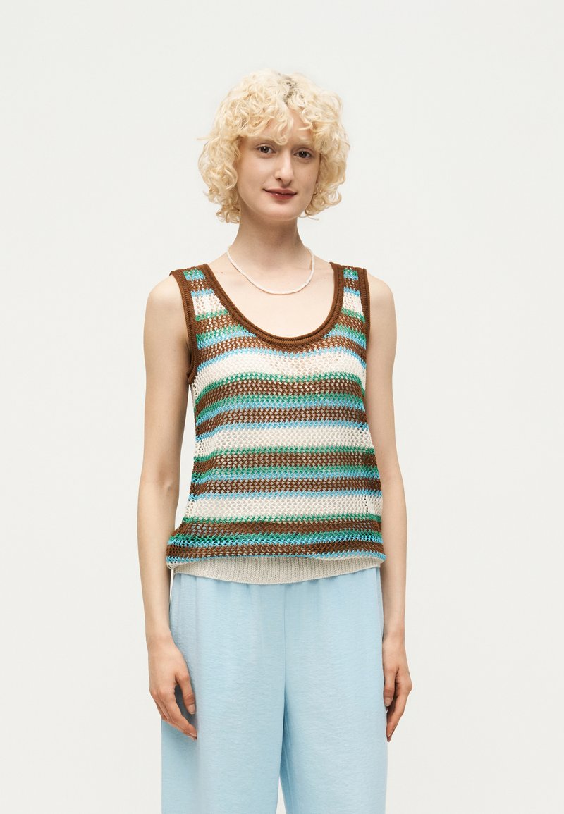 Jeune personne aux cheveux blonds bouclés portant un haut tricoté sans manches à rayures marron, bleu et crème, associé à un pantalon bleu clair.