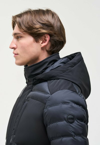 Un mannequin vu de profil porte une doudoune noire matelassée à capuche, avec un badge rond Colmar sur la manche.