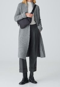 Larga cardigan gris texturizada con frente abierto, combinada con una blusa gris clara, pantalones gris oscuro y botas negras hasta el tobillo. Incluye bolso cruzado negro.