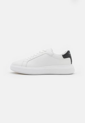Baskets basses blanches avec lacets blancs, semelle épaisse et languette de talon noire avec texte blanc partiel.