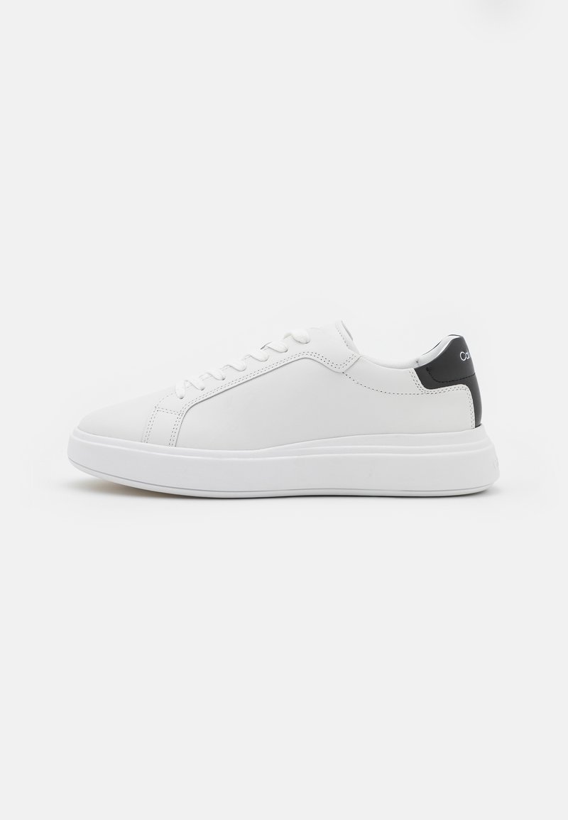 Baskets basses blanches avec lacets blancs, semelle épaisse et languette de talon noire avec texte blanc partiel.