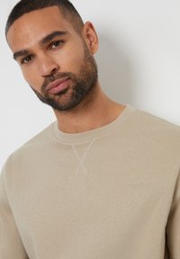 Sudadera beige con cuello redondo, presenta un detalle de punto de cruz en el escote y un pequeño logotipo bordado en el lado izquierdo del pecho.