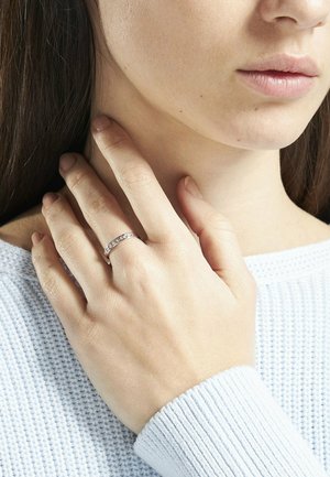Silberner Ring mit fünf kleinen klaren Steinen, getragen am Ringfinger einer Hand, die den Hals berührt. Der Pullover hat ein strukturiertes, hellblaues Strickmuster.