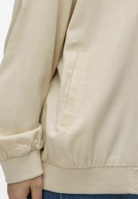 Beige jacket with a relaxed fit, elastic cuff, side pocket, and textured fabric. Features a banded hem.