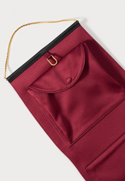 Viininpunainen kangaspussi kultaisella ketjusangalla, musta yläreuna, pyöreä läppä ja etutasku, joissa on painonappi ja koristeompeleet.