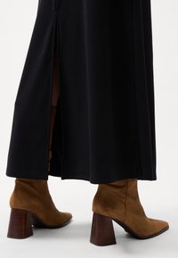 Robe longue en tissu noir avec une fente sur le côté, associée à des bottes en daim marron arrivant à la cheville, dotées d'un talon en bois épais.
