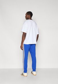 Biela nadrozmerná tričko s krátkymi rukávmi, skombinované s modrými teplákmi so žltými bočnými pruhmi. Čierne a biele tenisky dotvárajú vzhľad.